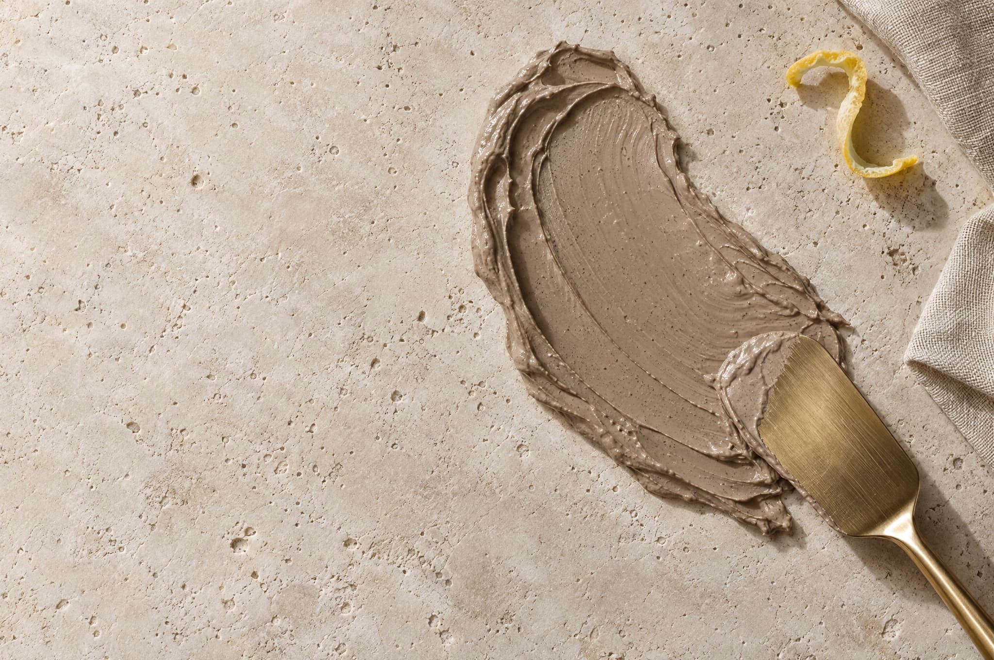 Taupe mud mask spread on stone with a brass spatula